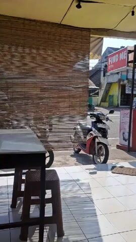 A motorcycle is parked outside a building. A sign that says "FIND ME" is visible. There are tables and chairs in the foreground. Sunlight casts shadows on the ground. A water jug is placed on a small stool. A tree is visible in the background.