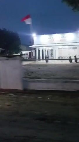 The video captures a dimly lit outdoor scene at dusk or night. In the background, a white building with illuminated windows and a sign above the entrance is visible. Several motorcycles are parked in front of the building. A fence runs along the foreground, and trees are present in the environment. The camera appears to be moving, giving a dynamic perspective of the location.