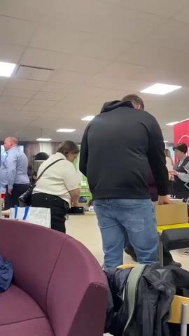 The video shows a man standing with his back to the camera, wearing a black hoodie and blue jeans, in what appears to be a public event space. In the background, several people are standing around tables and displays. A woman to the left is looking at something on a table, while another woman in a purple top stands by a red banner with the letters "BF" on it. A man in a white jacket holds a long, thin pole. The space has a low, white ceiling with fluorescent lights. A purple couch is visible in the foreground. There are boxes and other items on and around the tables. The atmosphere suggests a conference or exhibition setting.