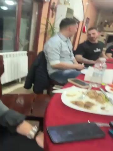 Several men are gathered around a long table covered with a red tablecloth, enjoying a meal. The table is laden with plates of food, water bottles, juice, and bottles of alcohol. Some of the men are talking, while others are looking at their phones. One man is using an electronic cigarette. The room appears to be a restaurant, with decorative elements and arched windows visible in the background. Music is playing in the background.