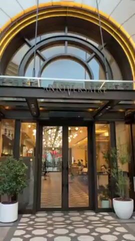 The video shows the exterior of the "Anniversaire Omotesando" building in Japan. The camera slowly zooms in on the building's entrance, which features glass double doors framed by a dark metal structure. Above the doors, the building's name is displayed in silver lettering. Through the glass, the interior appears to be a cafe or restaurant with patrons seated at tables. Decorations, including strings of lights, are visible inside. Outside the building, there is a glimpse of an outdoor area with people walking. The sky is overcast.