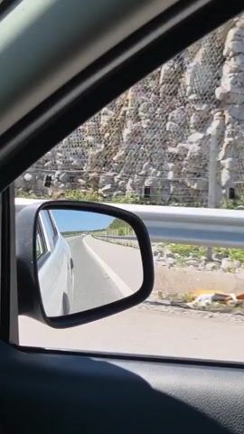 The video is filmed from inside a car looking out the passenger side window and into the side mirror. The car is in motion on a paved road. To the right of the road, there is a white metal guardrail. Beyond the guardrail, there is a rocky, steep hillside that has been reinforced with a mesh netting system. The sky appears clear and blue. The side mirror reflects the road behind the car, showing the white line dividing the lanes and the car's rear bumper. The overall scene suggests travel through a mountainous or hilly region.