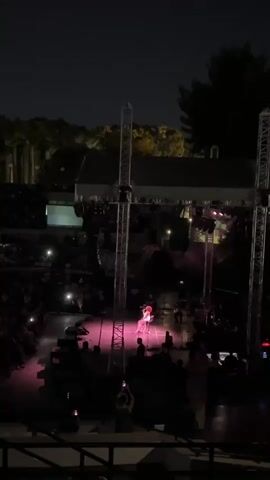 A concert is taking place at night on an outdoor stage. The stage is lit with pink lights, and there are several tall scaffolding structures on either side of the stage. A performer, wearing a red outfit, is visible on stage, standing and holding a microphone. A crowd of people is seated in an amphitheater in front of the stage, many of them holding up their phones, presumably to record or photograph the performance. Trees are visible in the background behind the stage.