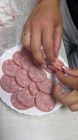 A pair of hands is shown close up, arranging sliced smoked sausage on a white plate. The sausage is pinkish-red with flecks of white fat and a darker rind around the edge. The plate is filled with evenly sliced sausage pieces, overlapping each other. One hand, with neatly manicured nails, is holding a slice of sausage and carefully placing it on the plate. A gold ring is visible on the index finger of the other hand, which is also holding sausage slices. The background is blurred, but appears to be a kitchen counter with a plastic bag and some gray fabric. The lighting is bright and focused on the hands and the sausage.