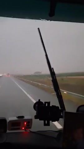 The video provides a first-person perspective from inside a vehicle driving on a highway during a rainstorm. The windshield is wet with rain, creating streaks and obscuring the view. The road ahead is wet and appears to have puddles. The sky is overcast and gray, indicating heavy rain. Orange traffic cones are visible on the side of the road, lining the shoulder. Other vehicles with illuminated headlights can be seen in the distance on the highway. Inside the vehicle, a device with a red light is visible on the dashboard, possibly a radar detector or other electronic equipment. A phone mount is also attached to the windshield. The overall environment suggests challenging driving conditions due to the weather.