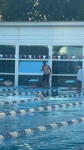This video shows an outdoor swimming pool with several people. In the foreground, the blue water of the pool is visible with lane lines. In the background, there is a building with large windows. Three people are standing by the pool. One person, a male, is shirtless and wearing patterned swim trunks. Another person, a female, is wearing a white long-sleeved shirt and blue shorts. A third person, also male, is standing to the left, partially submerged in the water. A fourth person, a female, is visible further to the left, also partially submerged in the water. A fifth person, a male, is swimming in one of the lanes. There are starting blocks at the edge of the pool, and colorful triangular flags are hanging above the windows of the building. A digital clock on the building shows the time as 19:56. The lighting suggests it is daytime, likely late afternoon or early evening. The setting appears to be a swimming facility, possibly a competition or training event.
