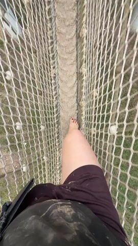 The video shows a first-person perspective of a person walking across a narrow rope bridge. The bridge is made of netting on the sides and a sandy-colored material in the center for walking. The person is barefoot and wearing dark shorts. The person's right foot is visible as they step forward. There are ropes and carabiners visible at the top of the bridge, indicating it is a suspended structure. The surrounding environment appears to be outdoors, with greenery visible below the bridge.