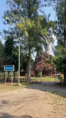 The video shows an outdoor scene with a dirt path leading towards a historical ruin. On the left side of the path, there is a blue sign with white text that reads "CANDI LOR". A tall street light is also visible to the left of the sign. The path is bordered by green grass and trees. In the background, there is a brick ruin partially overgrown with vegetation, with a large tree arching over it. A metal fence surrounds the ruin, and an open gate is visible in the foreground. The sky is blue with some clouds.