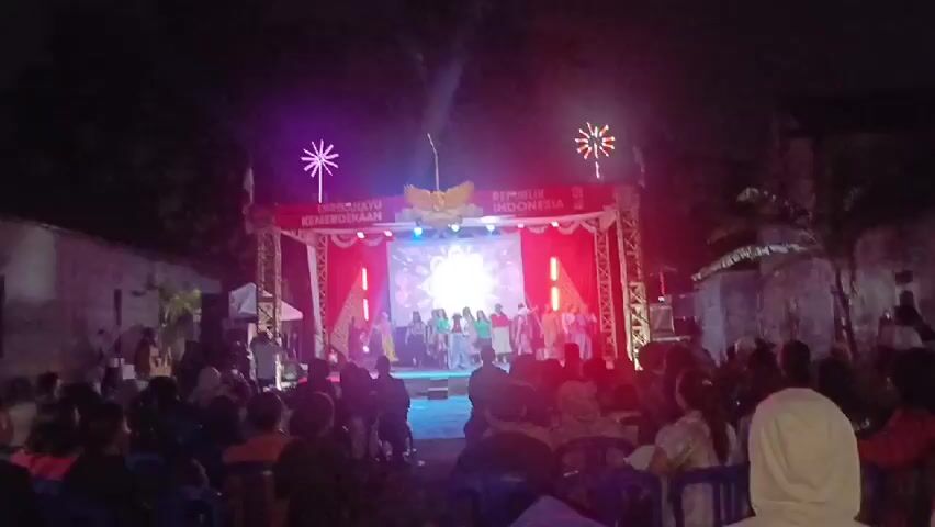A night-time outdoor stage event is depicted with a crowd of people seated in chairs watching a performance. The stage is illuminated with bright lights, featuring a backdrop with text and an emblem. A group of performers is visible on the stage. The scene suggests a celebratory or cultural gathering.