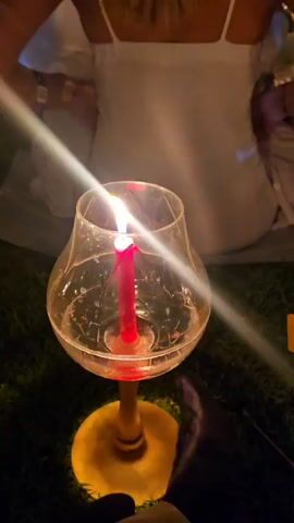 A woman in a white dress sits with others in a dark outdoor setting. Each person sits on the ground with a lit candle in front of them, the candles are in glass holders with water inside. In the distance, there is a large building, with a large screen visible above the heads of the people.
