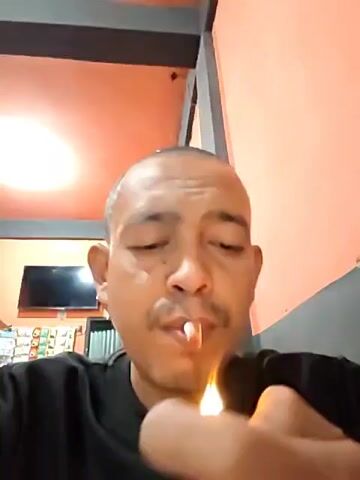 A close-up video shows a light-skinned man lighting a cigarette. He is wearing a black t-shirt and has a shaved head and a mustache. He lights the cigarette with a lighter in his right hand, then takes a puff and exhales smoke directly towards the camera. The background features a wall painted in a peachy hue and a drop ceiling. A television and snack display are visible in the background.