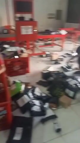 The video shows a busy warehouse or sorting facility. Several people, mostly wearing red shirts and some with caps, are engaged in sorting and organizing packages. The environment appears to be a large, open space with a tiled floor and fluorescent lighting. There are numerous red plastic crates scattered around, some filled with packages and others empty. Packages of various sizes and types, many in plastic wrapping or envelopes, are piled on the floor and inside the crates. In the background, there are conveyor belts and other sorting equipment, suggesting an organized operation. Some individuals are seen actively handling the packages, placing them into crates or moving them from one area to another. The overall impression is one of intense activity and movement within a logistics or distribution center.