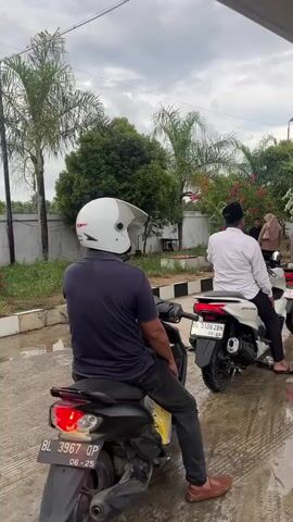 A line of motorcycles with riders is parked under a covered area, possibly a gas station. The sky is overcast. In the foreground, a man wearing a dark shirt and a white helmet sits on a black motorcycle. Further down the line, a man in a white shirt and black cap is on a white motorcycle. Other motorcycles and riders are visible behind them, stretching towards a service station attendant in a red uniform who stands near a fuel pump. The ground appears wet, suggesting recent rain. Trees and foliage are visible in the background.