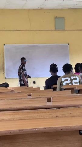 The video shows a small lecture hall, sparsely populated with students seated at wooden desks arranged in rows. A young man stands at the front of the room facing the students. He is writing on a whiteboard mounted on the yellow wall. The students are facing him, mostly from the back view. The room is brightly lit and the desks appear well-used with visible wear. The students are wearing a variety of clothes, and their posture suggests that they are paying attention to the lecturer. The atmosphere is typical of a classroom setting, conducive to learning. A utility box is mounted above the whiteboard.