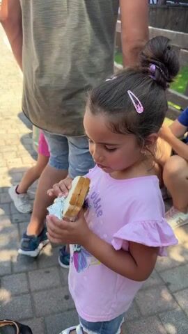 The video shows a young girl in a pink shirt and denim shorts with her hair in a bun eating a sandwich. She is holding the sandwich with both hands, and it appears to be wrapped in foil. She is standing outdoors on a paved pathway, and there are other people in the background, including a person in a green shirt and denim shorts. In the distance, there is an amusement park with rides and attractions, including a large white structure that resembles a flying saucer. The weather is sunny.