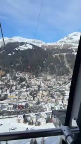 The video shows an aerial view of a snow-covered town from inside a cable car.  Mountains are visible in the background under a clear blue sky. The town consists of buildings covered in snow, and trees populate the mountainside behind it.  The cable car wires are visible in the foreground. The cable car is in motion.