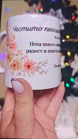 The video shows a close-up of a white ceramic mug held in a person's hand. The mug has a floral design on the left side and text printed on it. The text reads "Честито пенсиониране, Ваня!" (Happy retirement, Vanya!) at the top, followed by a date "16.12.2025". Below that, it says "Нека новото начало ти донесе радост и повече време за себе си" (May the new beginning bring you joy and more time for yourself). At the bottom, it is signed "От Крем`и" (From Kremy). The background is blurred, but appears to be a decorated Christmas tree with lights. The person holding the mug has manicured fingernails, with the ring finger and pinky finger visible, painted with white polish and glitter.