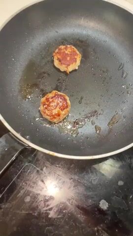 The video shows two small, round, cooked food items sizzling in a black non-stick frying pan with oil. The food items appear to be some kind of patty or fritter, with browned exteriors and visible texture. The pan is on a black stovetop. In the next sequence, a silver pot filled with boiling water is shown. Yellowish pasta is being stirred in the boiling water with a fork. The pot is also on a black stovetop.