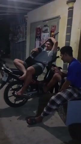 A man is sitting on a motorcycle holding a guitar. Another man is sitting next to him and looking at him. The motorcycle is parked on a paved surface in front of a building. The lighting suggests it is evening or night. The man on the motorcycle is wearing a grey t-shirt and shorts. The man sitting next to him is wearing a blue t-shirt and plaid pants.