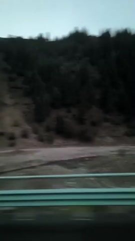 The video shows a mountainous landscape with a dark forest covering the upper slopes. A dirt road or path is visible on the lower part of the slope, with a light-colored guardrail in the foreground. The sky is overcast and appears to be twilight. The video is shaky, suggesting it was filmed from a moving vehicle.