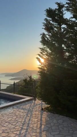 The video shows a sunrise over a coastal landscape, framed by a large evergreen tree on the right. A portion of a swimming pool is visible on the left, surrounded by a stone patio. A black metal fence separates the pool area from the vista, which includes mountains and the sea under a clear, blue sky. The sun is low on the horizon, casting long shadows from the tree onto the patio.