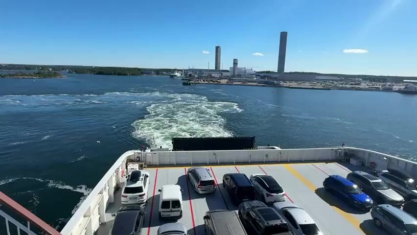 A ferry moves away from a dockyard towards open water. The ferry is carrying cars on its deck. A wake trails behind the ferry in the water. In the background, there are industrial buildings and smoke stacks. The sky is blue and clear with a few wispy clouds.