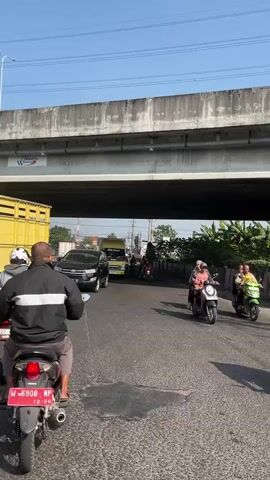 The video shows a busy street scene under an elevated concrete structure, possibly a bridge or overpass. The sky is clear and blue, suggesting daytime. The road is asphalt, with some visible patches and wear.

In the foreground, a person on a motorcycle is facing away from the camera, looking towards traffic. The motorcycle has a license plate that reads "88 NP" and a sticker with dates. The rider is wearing a black jacket with white stripes on the sleeves and gray shorts.

Further down the road, several vehicles are moving. A black SUV is in the left lane, and a yellow truck with a closed cargo box is in the right lane, moving away from the camera. Several motorcycles are also present. One motorcycle in the right lane carries two passengers, a driver and a passenger in the back. Another motorcycle further ahead has one rider.

On the right side of the road, there's a roadside with foliage, including what appears to be banana trees. There are also signs or structures visible along the right edge of the road. The elevated structure above is gray and has a rough concrete texture. Some cabling or wires are visible running along the underside of the structure. The overall impression is a typical urban or suburban road with moderate traffic flow.
