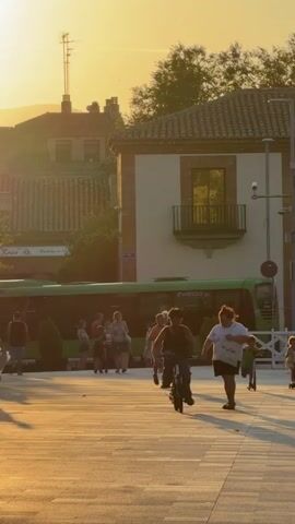 The video depicts a sunlit outdoor scene, likely a town square or pedestrian area, with a warm, golden hue cast by the low sun. In the foreground, several people are engaged in various activities. A young person on a bicycle is pedaling towards the camera. To their right, another young person is riding a scooter, also moving in the same direction. Behind them, two adults are walking, one with their back to the camera, and another facing slightly towards the left. Further to the left, a person is standing with their back to the camera, and several other individuals are visible in the background, including some walking near a green bus. The environment includes buildings with terracotta roofs and white facades, trees, and what appears to be street furniture and temporary barriers. The overall impression is one of a relaxed, public gathering with people enjoying the late afternoon or early evening light.