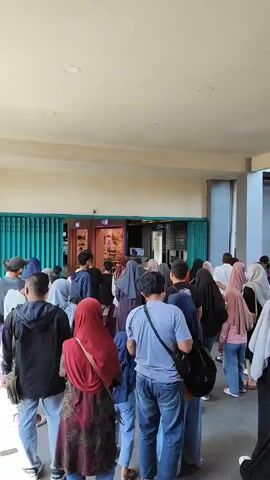 The video shows a long queue of people standing in what appears to be an indoor or covered outdoor walkway. They are facing a shop with a green metal gate-like facade. The people are diverse in age and gender, with many wearing headscarves. They are standing in line, facing the shop, with some individuals closer to the shop entrance than others. The environment seems to be a public space, possibly a mall or a shopping complex, with a light-colored ceiling and columns visible. The people are mostly stationary, indicating they are waiting.