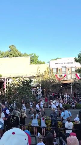 The video shows an outdoor event with a crowd of people gathered. In the background, there are buildings, one of which appears to be a historical museum with "FOLSOM HISTORY MUSEUM" signage. Red, white, and blue bunting is draped across the front of the museum. Several trees with green foliage are prominent in the scene, some taller than the buildings. A group of people are visible in the foreground, facing away from the camera, and appear to be watching something or someone in the distance. Some individuals are holding cell phones, possibly recording the event. The overall atmosphere suggests a public gathering or festival under a clear blue sky.