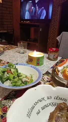 A close-up shot of a table set for a meal. In the foreground, a white bowl with dark text appears to contain meat. To the left, a blue and white bowl holds a salad with lettuce, cucumber, and tomato. A lit yellow candle sits between the bowls, with text visible on its label. A glass of water is behind the candle, and a jar of mustard is to the right. In the background, a television screen displays a scene from a movie featuring two people. The table is covered with a floral-patterned tablecloth.