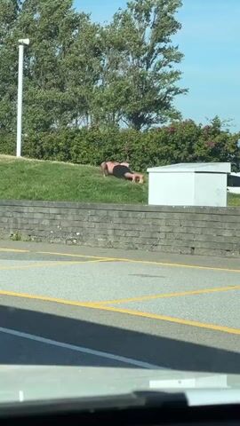 The video captures a shirtless individual performing push-ups on a grassy incline. The person is positioned facing away from the camera, with their body in a plank position. They are dressed in dark shorts. Behind them, a white rectangular structure and a low stone wall are visible. The background features trees and a clear blue sky. The scene appears to be outdoors, possibly near a parking lot, as indicated by the yellow lines on the ground in the foreground. The lighting suggests it is daytime.
