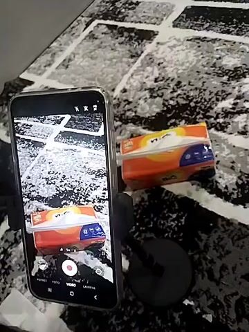 A smartphone, mounted on a small black stand, is recording a pack of tissues on a rug. The tissues are in an orange and blue wrapper that reads "Nice". A single tissue is partially pulled out from the pack. The rug has a black and white patterned design. The smartphone screen displays a live view of the same scene it is recording. The camera is positioned in such a way that it captures both the tissue pack and the smartphone simultaneously. The environment appears to be indoors.