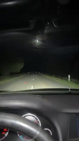 The video shows a first-person perspective from inside a car driving on a road at night. The headlights illuminate the road ahead, and the moon is visible in the dark sky. The dashboard and steering wheel are partially visible in the foreground. There are streetlights or other lights visible in the distance along the road.
