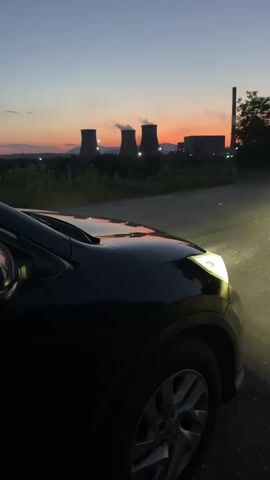 A black car is parked on the side of a road at dusk, with its headlights on. In the background, several cooling towers from a power plant are visible, with steam or smoke coming from the tops. The sky is a mix of deep blue and orange from the sunset. The overall scene suggests a moment of quiet observation of industrial landscape at the end of the day.