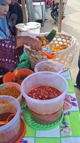 A woman wearing a blue hijab and a patterned dress is serving food from a street stall. She is using an orange ladle to place food from a large woven basket onto a plate. Several containers of food are visible on the table in front of her, including a large clear plastic tub filled with red stew-like food containing cubed ingredients, another clear plastic tub with a dark red sauce and a ladle, and an orange bowl with shredded reddish-brown food and green chopped ingredients. A young person stands behind her, looking at the food. The street stall appears to be set up outdoors, with a road and other stalls visible in the background.