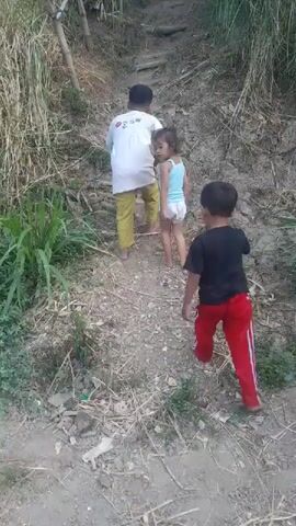 Three children are walking up a steep, dirt path. The child in the front is wearing a black t-shirt and red pants. The child in the middle is wearing a white shirt and yellow pants. The child in the back is wearing a light blue shirt and white shorts. The path is lined with dry grass and some green plants. There are some bamboo poles on the left side of the path. The ground is uneven and rocky. The overall impression is one of a rural, outdoor setting with children engaging in physical activity.