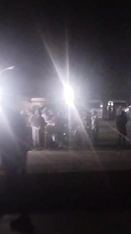 The video shows a dark outdoor scene with a group of people gathered around illuminated by bright lights, possibly at a gas station or similar establishment, with cars parked nearby. The focus is not sharp, making it difficult to discern specific details. People are standing and appear to be interacting with each other, but the exact nature of their actions is unclear due to the image quality.