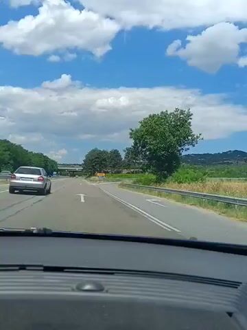 The video shows a first-person perspective from inside a car driving on a highway. The sky is blue with scattered white clouds. Trees and greenery line the sides of the road. There is a silver car in front of the vehicle, traveling in the same direction. Ahead, there is a junction or exit with road signs. The road is asphalt, and there are lane markings. The dashboard of the car is visible in the foreground. The weather appears to be sunny.