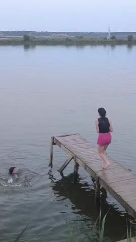 A woman in a life vest and pink shorts jumps off a wooden pier into a lake. Another woman is already in the water, swimming. The jumping woman makes a large splash. The water is calm and the sky is overcast. Trees and hills are visible in the background.