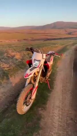 The video begins with a close-up of a white and red Husqvarna motorcycle parked on a dirt track in a field. The background shows rolling hills under a clear sky at dusk. The camera then pans to the right, showing more of the field with dry grass and sparse vegetation. The sun is setting on the horizon, casting a golden light. The sky transitions from blue to orange near the horizon. The video ends with a view of the open field at sunset.