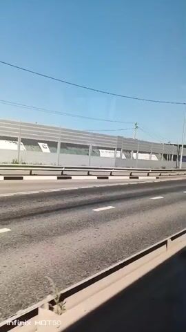 The video shows a view from a moving vehicle on a highway. The road has multiple lanes marked with white lines. On the left side of the road, there is a sound barrier, which appears to be made of metal panels. Behind the sound barrier, there are some buildings and trees visible. The sky is clear and blue. Several vehicles are seen on the highway, including a blue car, a silver van, and a black SUV. The camera is positioned to capture the passing scenery, suggesting the vehicle is in motion, traveling forward. The footage includes a watermark at the bottom that reads "Infinix HOT 50".