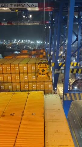 The video captures a night-time view of a busy container terminal. A large yellow gantry crane is actively engaged in loading or unloading shipping containers from a ship. The crane's mechanism is gripping a stack of orange containers. The ship itself is laden with numerous similar orange containers, stacked high. In the background, the port area is illuminated by numerous lights, indicating ongoing operations. A sign that reads "ABIDJAN TERMINAL" is visible on the gantry crane. The environment appears to be a typical industrial port setting with large machinery and vast quantities of goods.