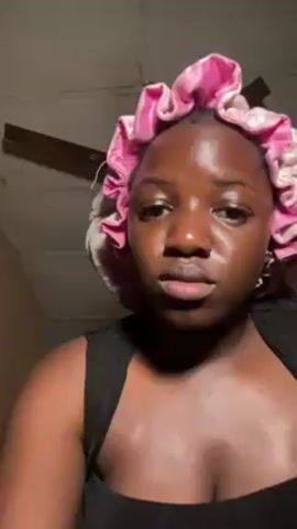 The video shows a woman applying lip liner to her upper lip with a makeup brush. The woman is wearing a pink ruffled bonnet and a black tank top. She appears to be in a room with a ceiling fan. The lighting is somewhat dim.