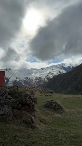 The video shows a mountainous landscape on a cloudy day. Snow-capped peaks stretch across the horizon, partially obscured by low-hanging clouds. In the foreground, a green field dotted with rocks leads towards the mountains. The sun is visible through a break in the clouds, casting a diffused light over the scene. A red building is partially visible on the left side of the frame. The camera pans slowly across the landscape.