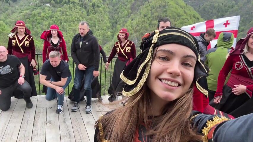 A young woman with long brown hair and blue eyes, wearing a traditional Georgian outfit, holds a camera to take a selfie with a group of people.  The group stands on a wooden deck with a railing, with a lush green mountain backdrop. Many of the people in the background wear traditional Georgian clothing, while others are in casual modern attire.  A Georgian flag is also visible in the background. The woman smiles directly at the camera, and the image is generally well-lit, suggesting an outdoor setting during the daytime. At one point a woman in a red outfit joins the selfie.