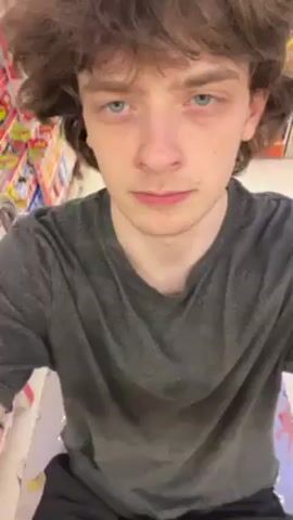 A young, white-skinned man with wavy brown hair and blue eyes films himself while shopping in a supermarket. He wears a grey t-shirt and black pants. He is standing behind a shopping cart, which is partially visible. The cart is moving through the aisles of the supermarket. Various products like sweets and canned goods are visible on the shelves around him. The camera is handheld, resulting in slight movements. The lighting in the supermarket appears to be bright and even.