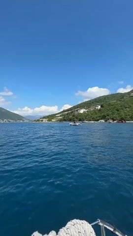 The video captures a serene coastal scene on a bright, sunny day. Filmed from a moving boat, the viewer sees calm, deep blue waters with gentle ripples. In the foreground, parts of the boat are visible, including a white rope coiled on the bow and a metal ladder on the side.

The shoreline is characterized by a lush green, forested hill dotted with several white and light-colored houses, suggesting a residential or resort area. A small, white motorboat is visible cruising on the water in the mid-ground. Further along the coastline, a partially submerged shipwreck with rusted red elements can be seen resting near the shore.

The sky is a clear, vibrant blue with a few scattered white cumulus clouds, indicating excellent weather conditions. The overall impression is one of a peaceful and picturesque maritime environment. The location is Luštica, Montenegro.