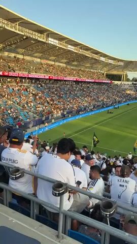 The video shows a wide shot of a soccer stadium from the perspective of the stands. The stands are filled with spectators, most of whom are wearing white jerseys. The field is green, with white lines marking the boundaries of play. The sky is clear and blue. A large screen is visible in the distance, displaying images of the game. A sign in the foreground reads "LA GALAXY SUPPORTER SECTION". Several people are visible in the foreground, facing away from the camera, watching the game. The atmosphere appears to be festive and energetic.