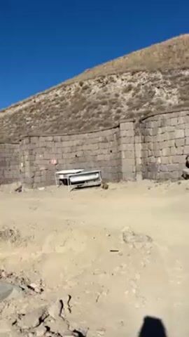 The video shows a dog lying in the shade of two old bathtubs in front of a stone wall with a grassy hillside behind it. A red-and-white no-entry sign is affixed to the wall. The area is a sandy-colored dirt area. A cellphone rings.